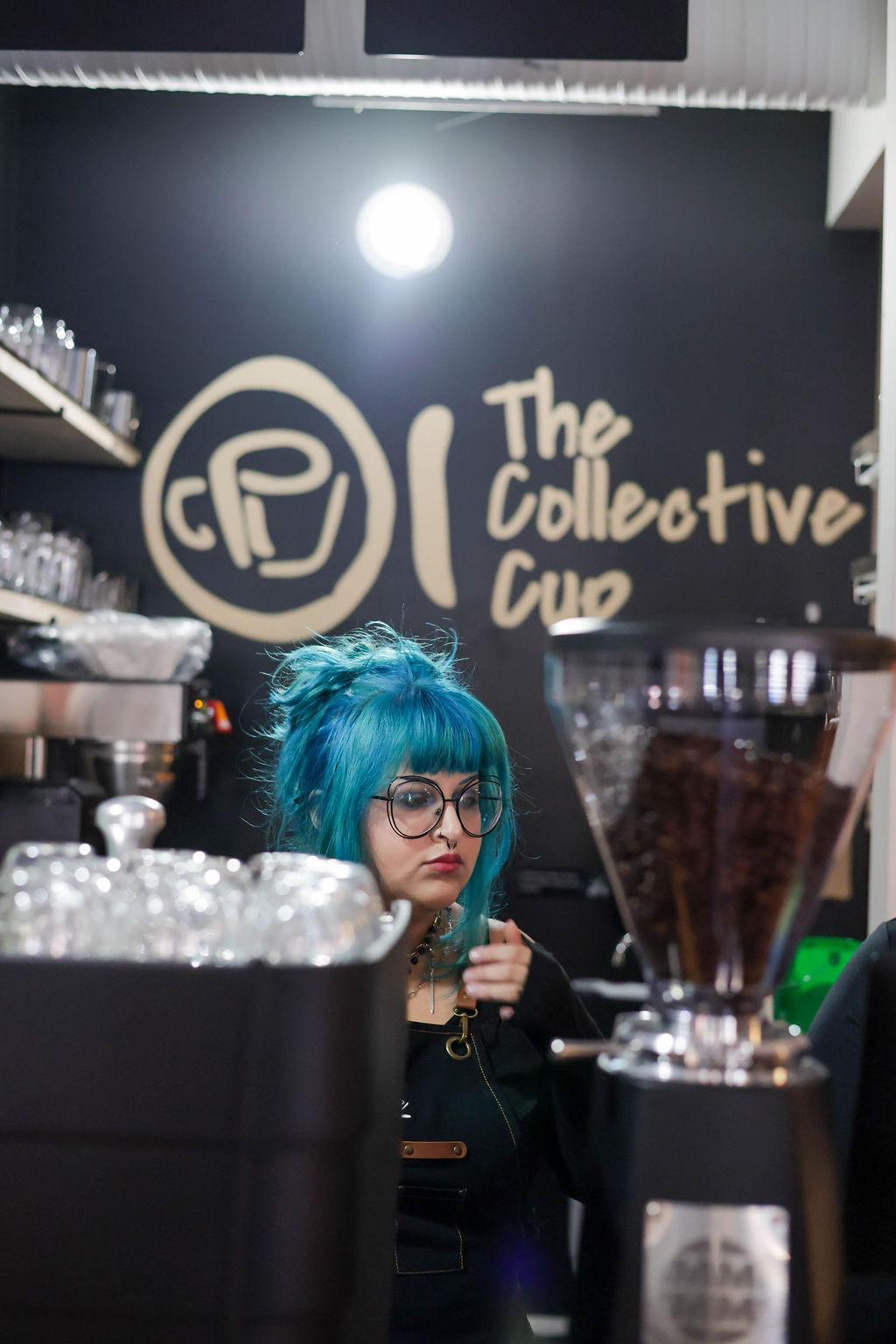 A barista with blue hair working behind the espresso bar beneath The Collective Cup lettering